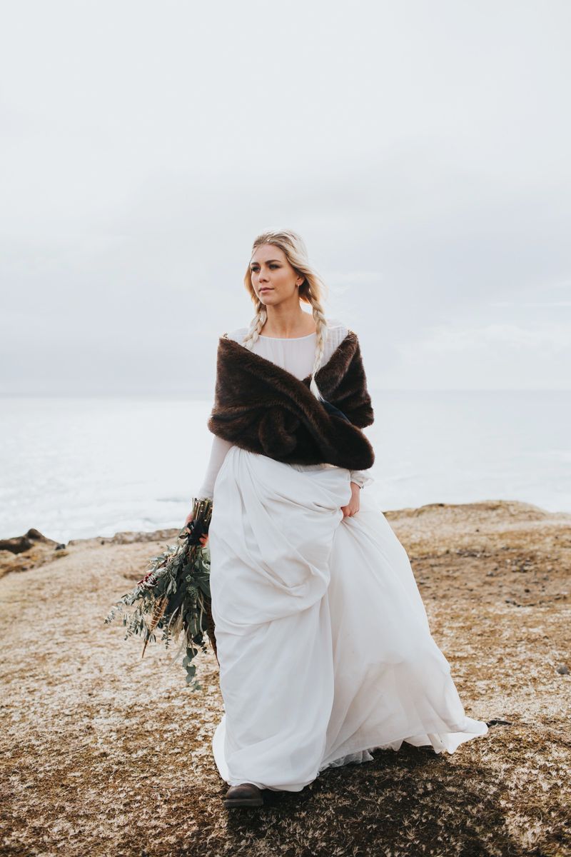 Bride in elegant white gown and fur stole poses on windswept clifftop.
