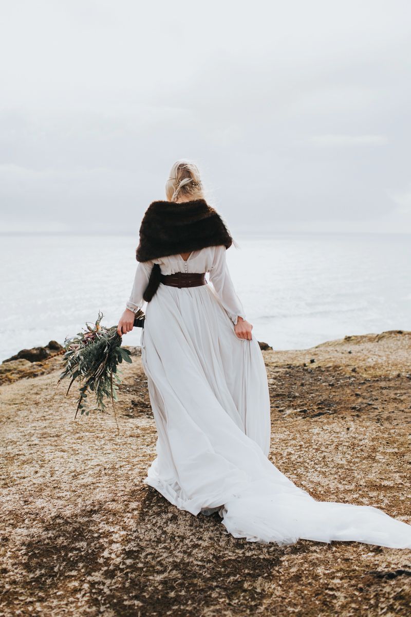 Flowing white wedding dress trails behind bride walking along rocky coastal path.