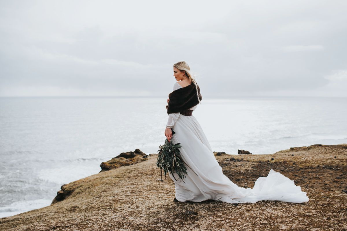 Bride stands on dramatic ocean cliff edge in flowing dress with winter bouquet.