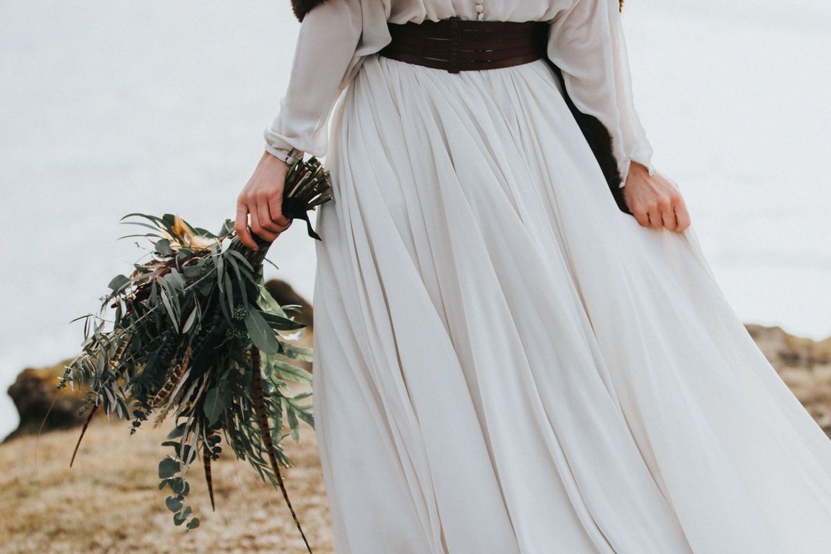 A flowing white wedding dress with black sash shown from waist down holding greenery bouquet against natural backdrop.