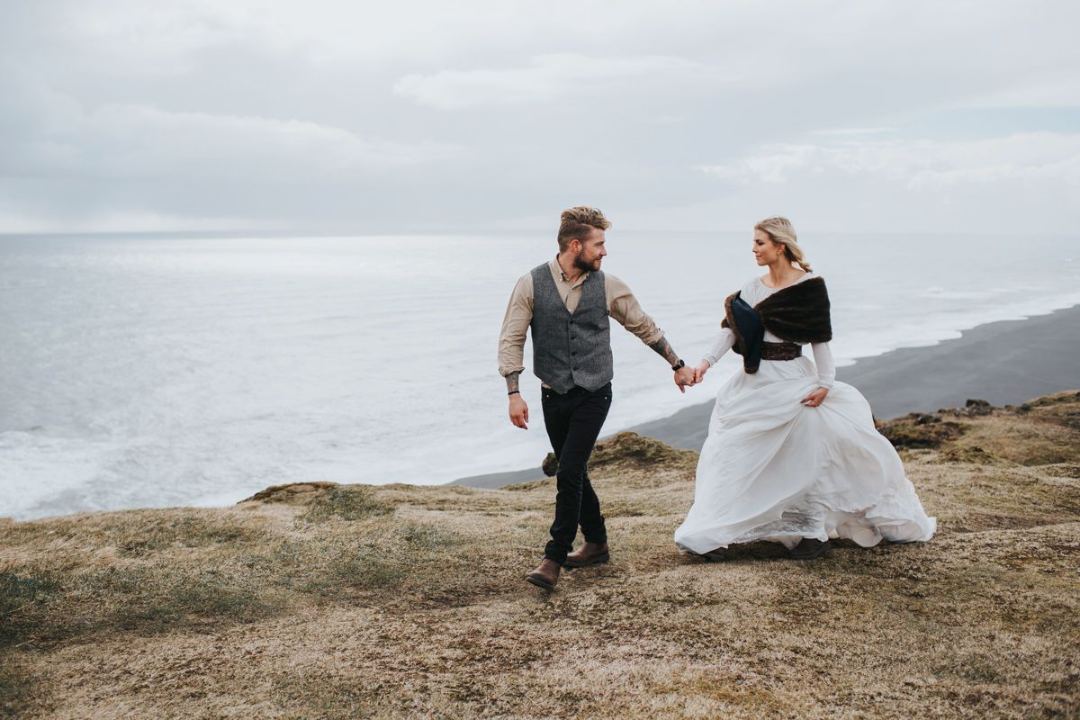 Wedding couple strolling hand in hand on rugged coastal cliffside with ocean view.