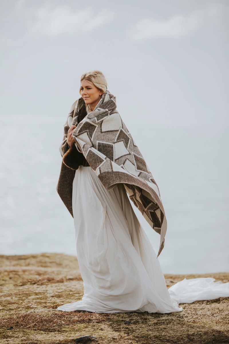 Person wrapped in geometric patterned blanket wearing white dress on coastal cliff.