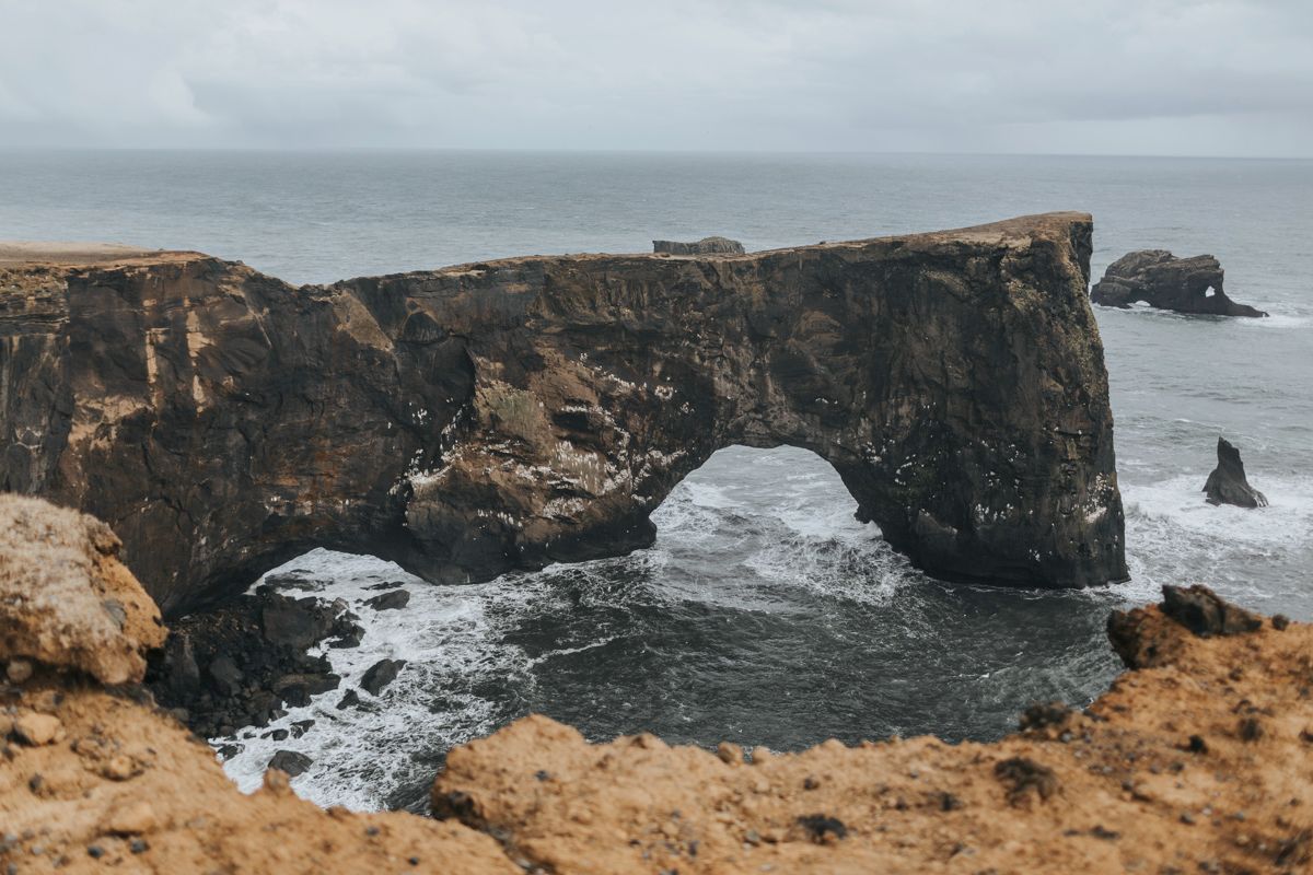 Dramatic rock arch formation on rugged coastline with crashing waves below.