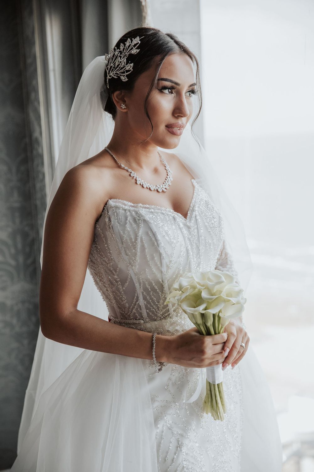 Elegant bride in lace gown with crystal hair accessory holds white calla lily bouquet by window.