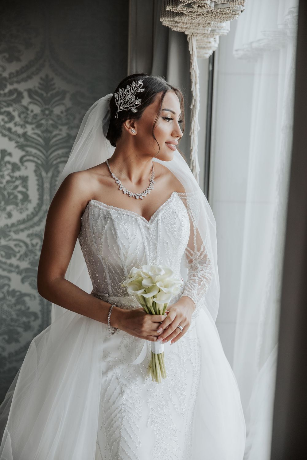 Bride in detailed lace wedding dress poses by window with crystal chandelier and damask wallpaper.