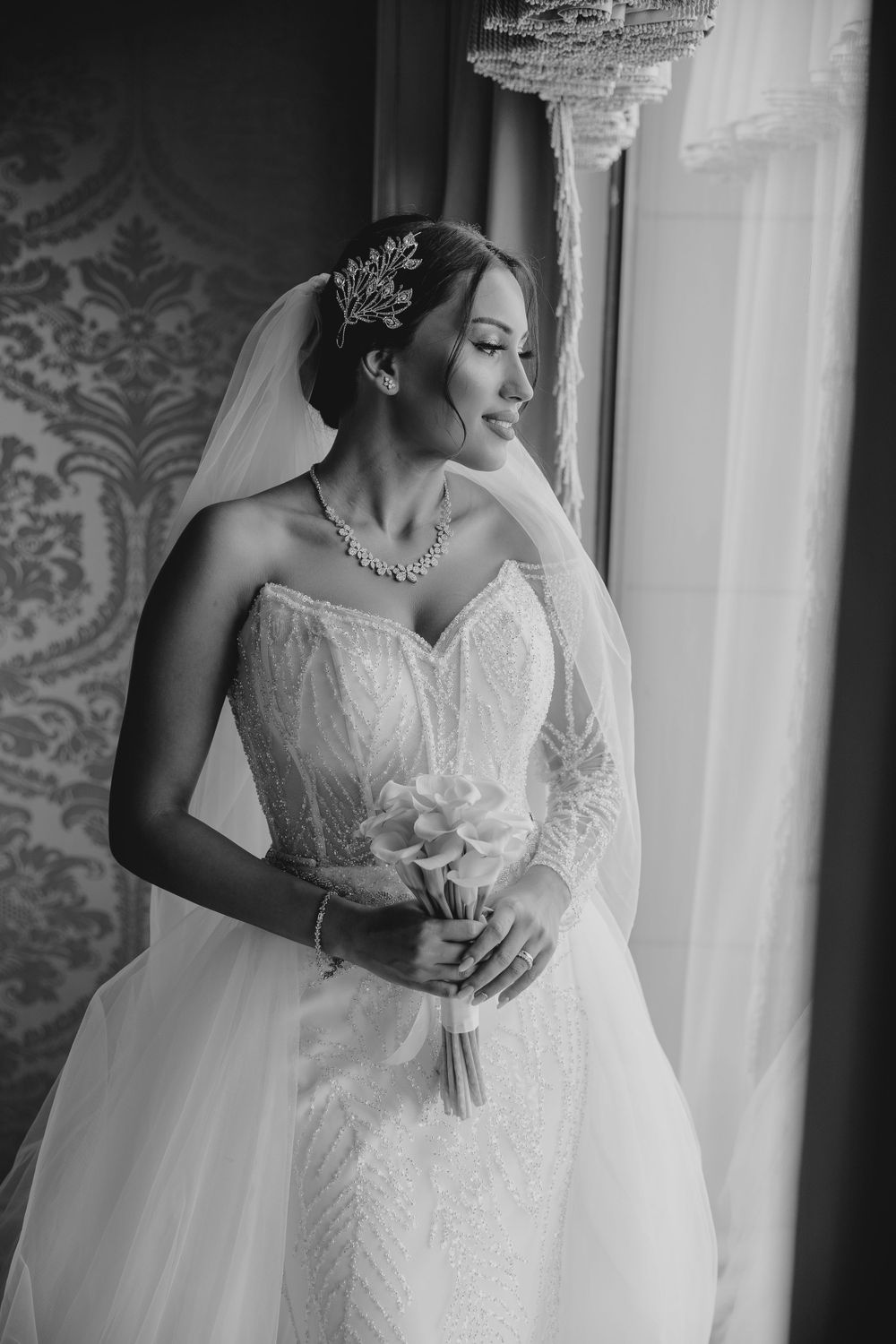 Black and white portrait of bride in strapless gown holding bouquet near ornate wallpaper.