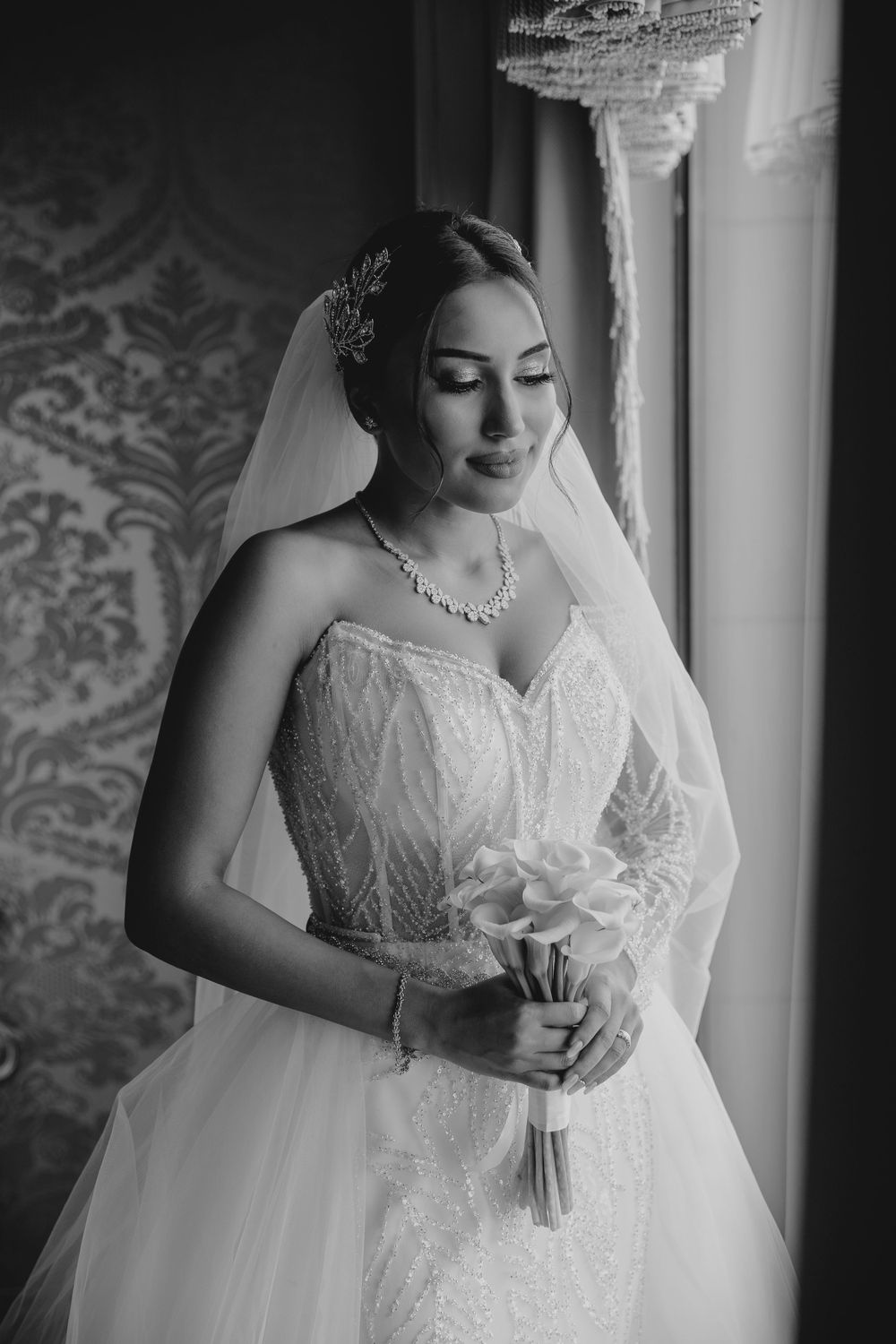 Monochrome bridal portrait showing intricate lace dress and crystal accessories near window.