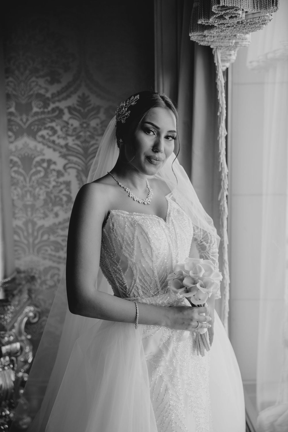 Black and white bridal photo with elegant jewelry and traditional veil by window.