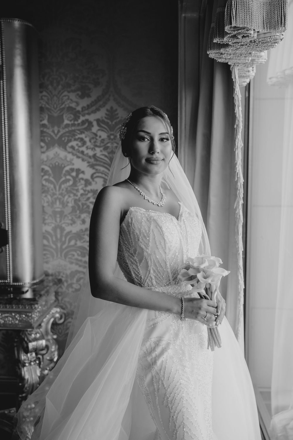 Black and white portrait of a bride in a strapless gown holding a white bouquet near a window.
