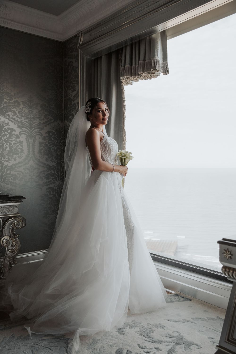 Ethereal wedding photo of a bride in flowing white gown against a bright window view.