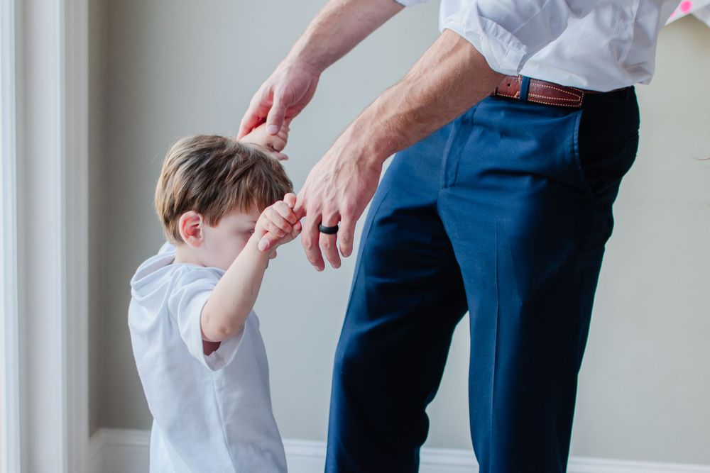 A toddler boy holds his dad's fingers as they dance in the kitchen.