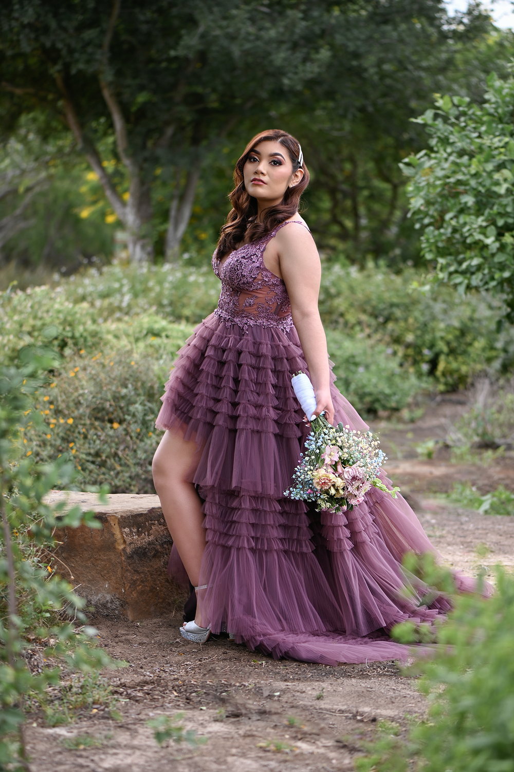 Purple formal dress with layered tulle skirt and bouquet photographed on stone bench in garden.