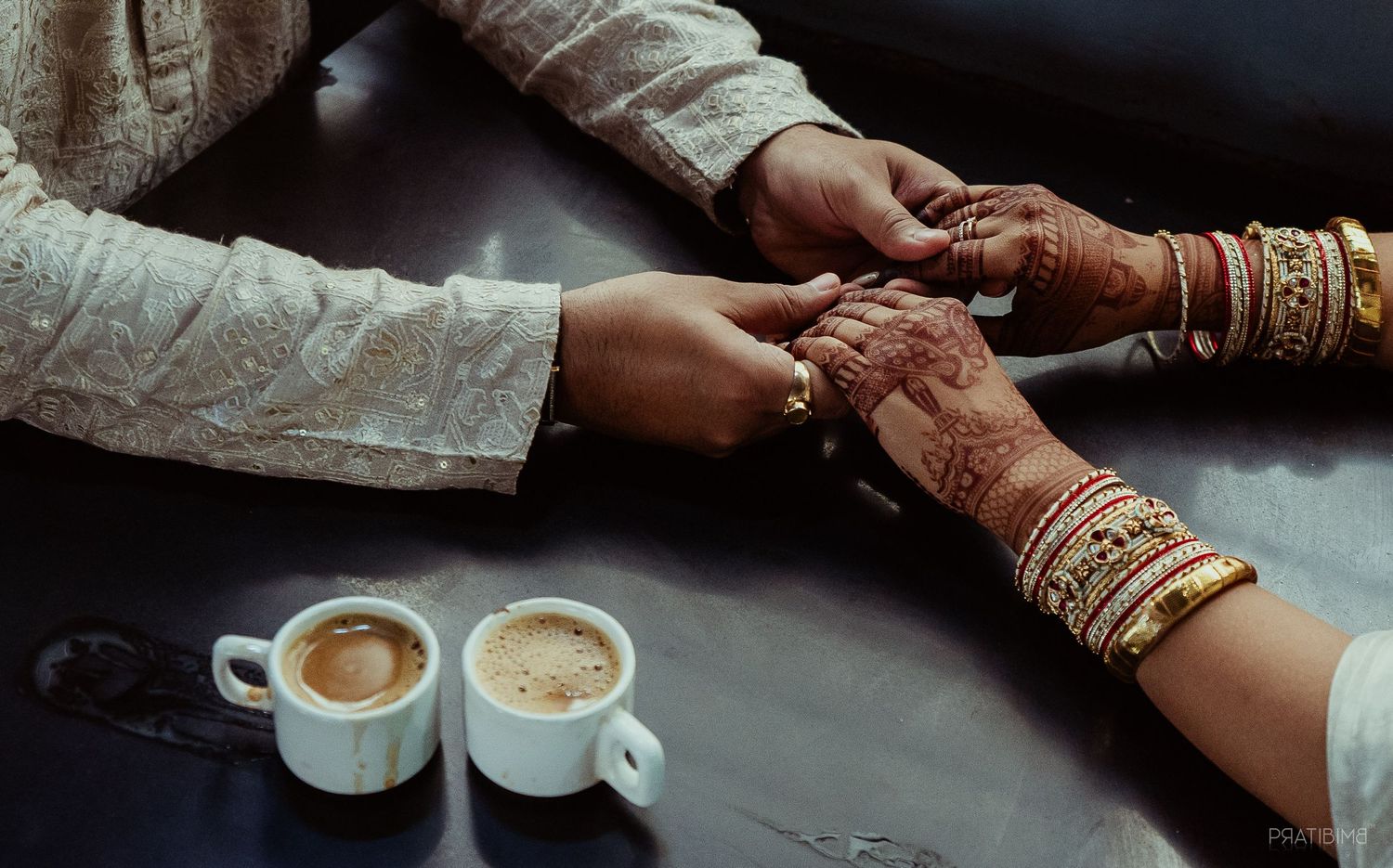 Close-up of hands with traditional bangles sharing coffee cups on dark table.