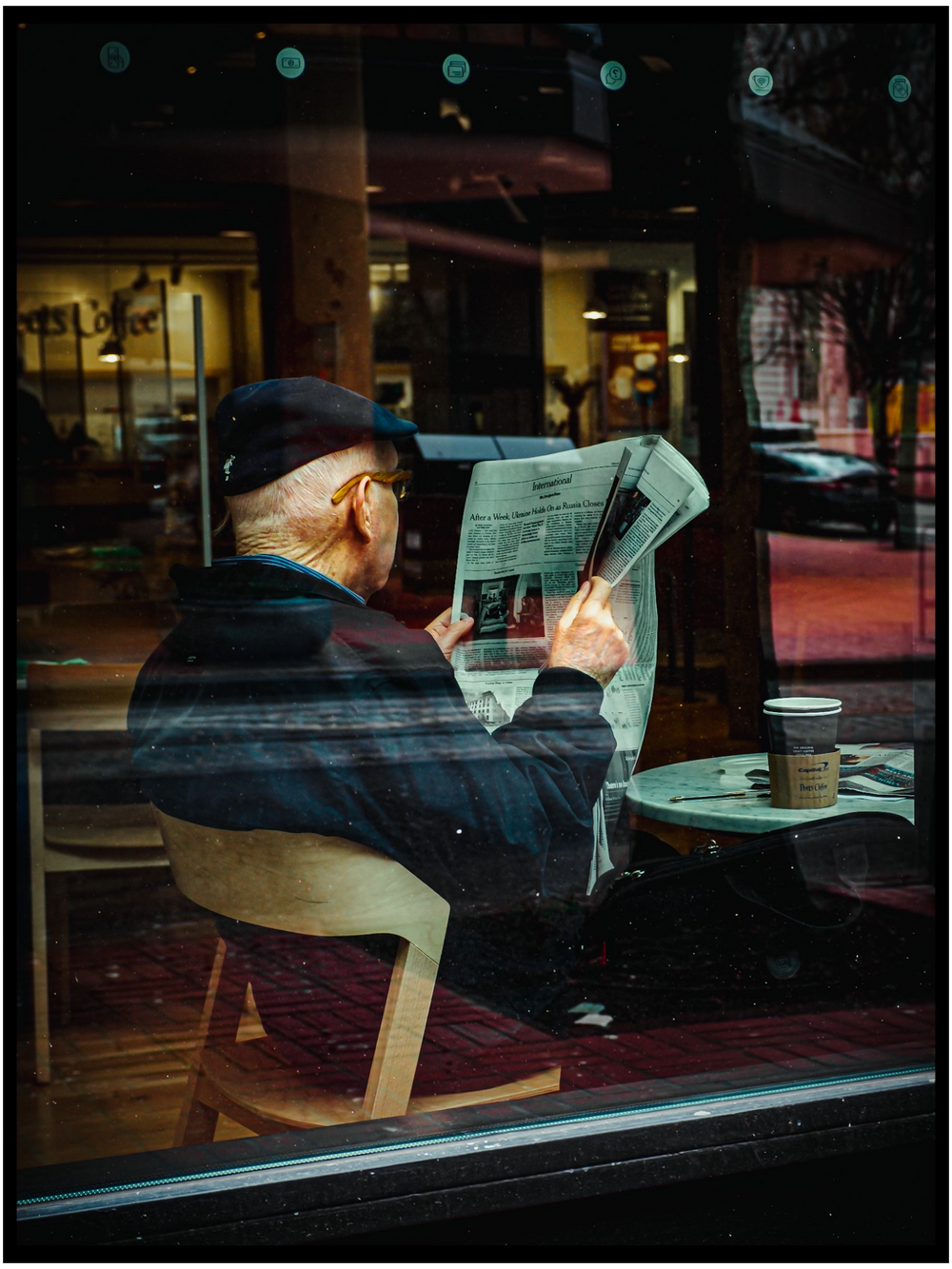 Elderly person reading newspaper by window in coffee shop with cup on table. Portland street photography