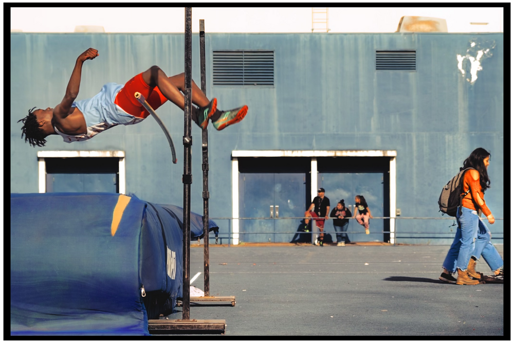 Person performing a high jump over blue equipment while others watch in an urban setting.