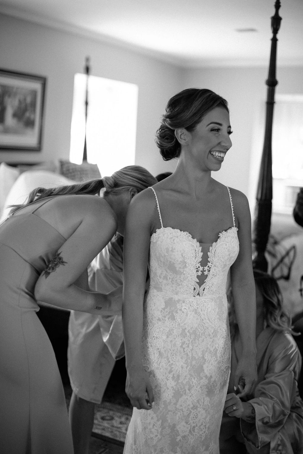A bride in an elegant lace wedding dress smiles while being fitted on her wedding day.