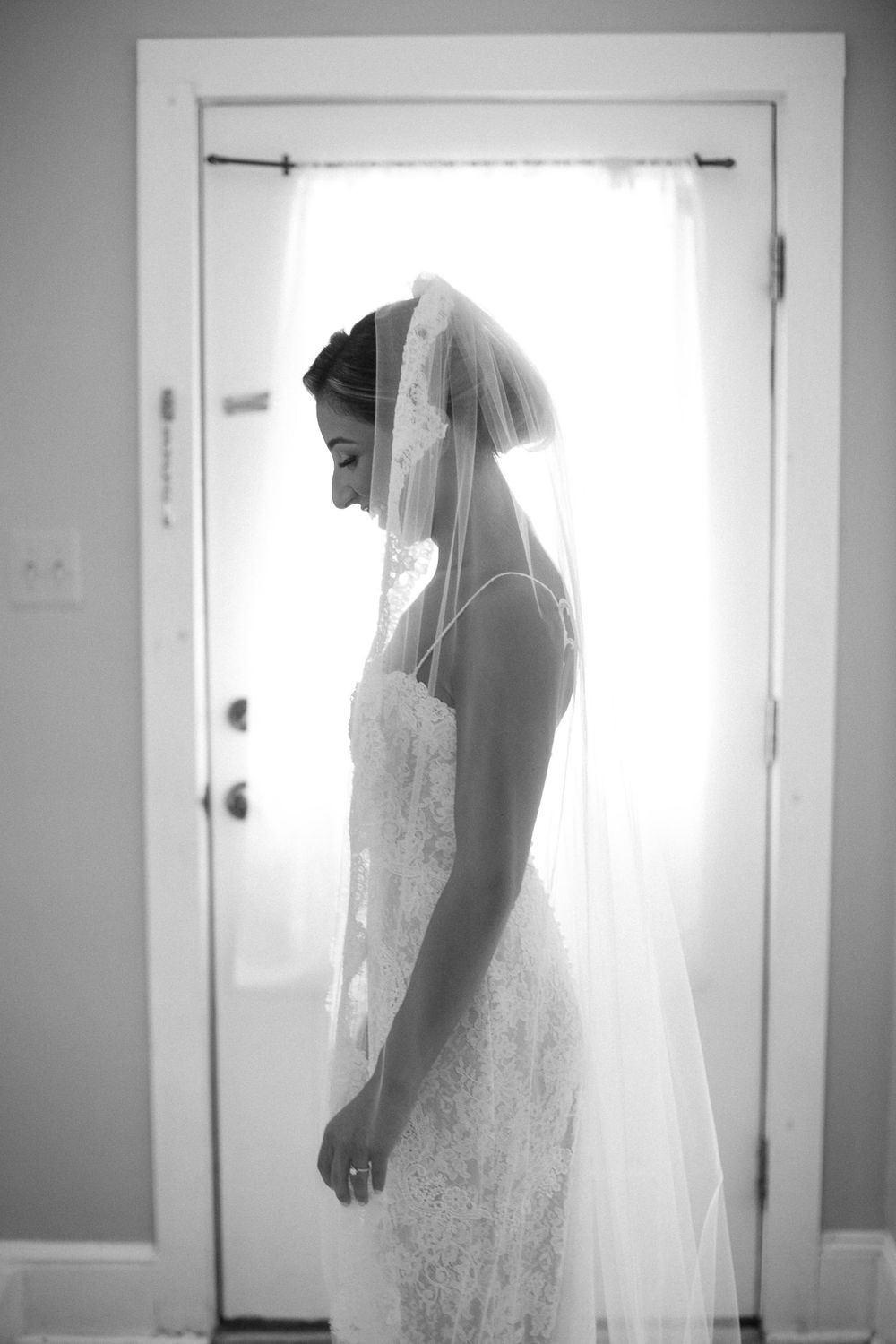 A silhouetted side view of a bride in a lace wedding dress and veil standing in front of a bright doorway.