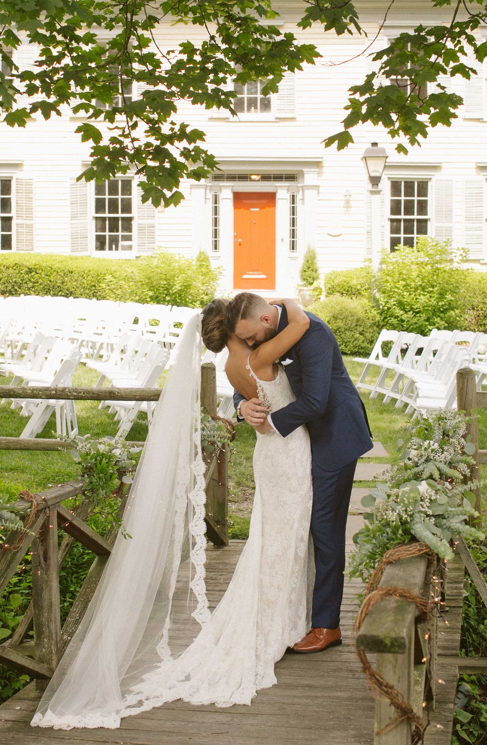 Newlyweds kiss on a wooden bridge with white colonial house and garden ceremony setup visible.