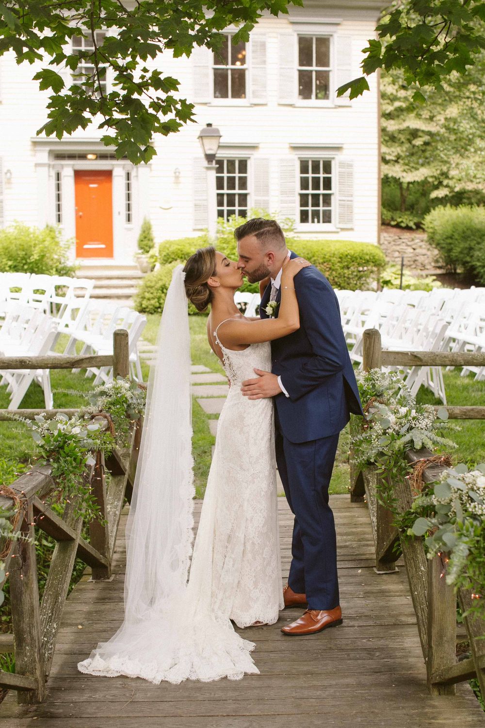 Wedding couple embracing on wooden bridge with white house and garden ceremony setup in background.