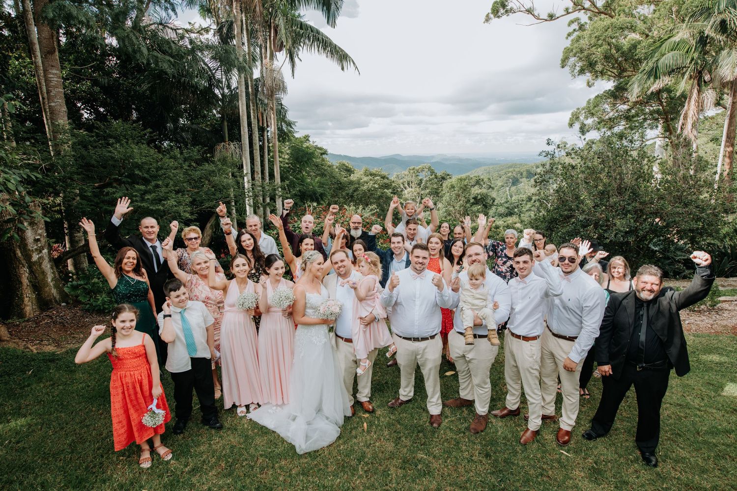 A large wedding party poses together on a hilltop with palm trees and mountains in the background.