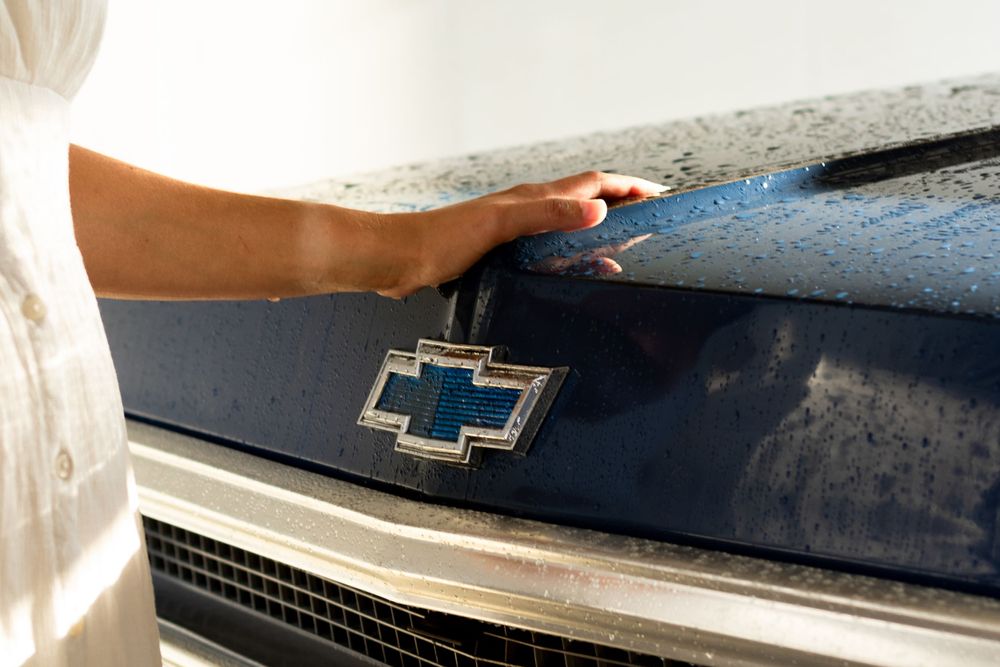 Hand touching the Chevrolet bowtie emblem on a dark blue vintage car.