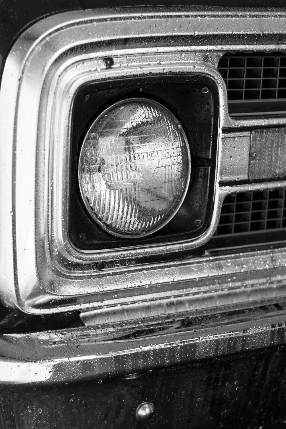 Close-up black and white photo of vintage car headlight and grille detail.