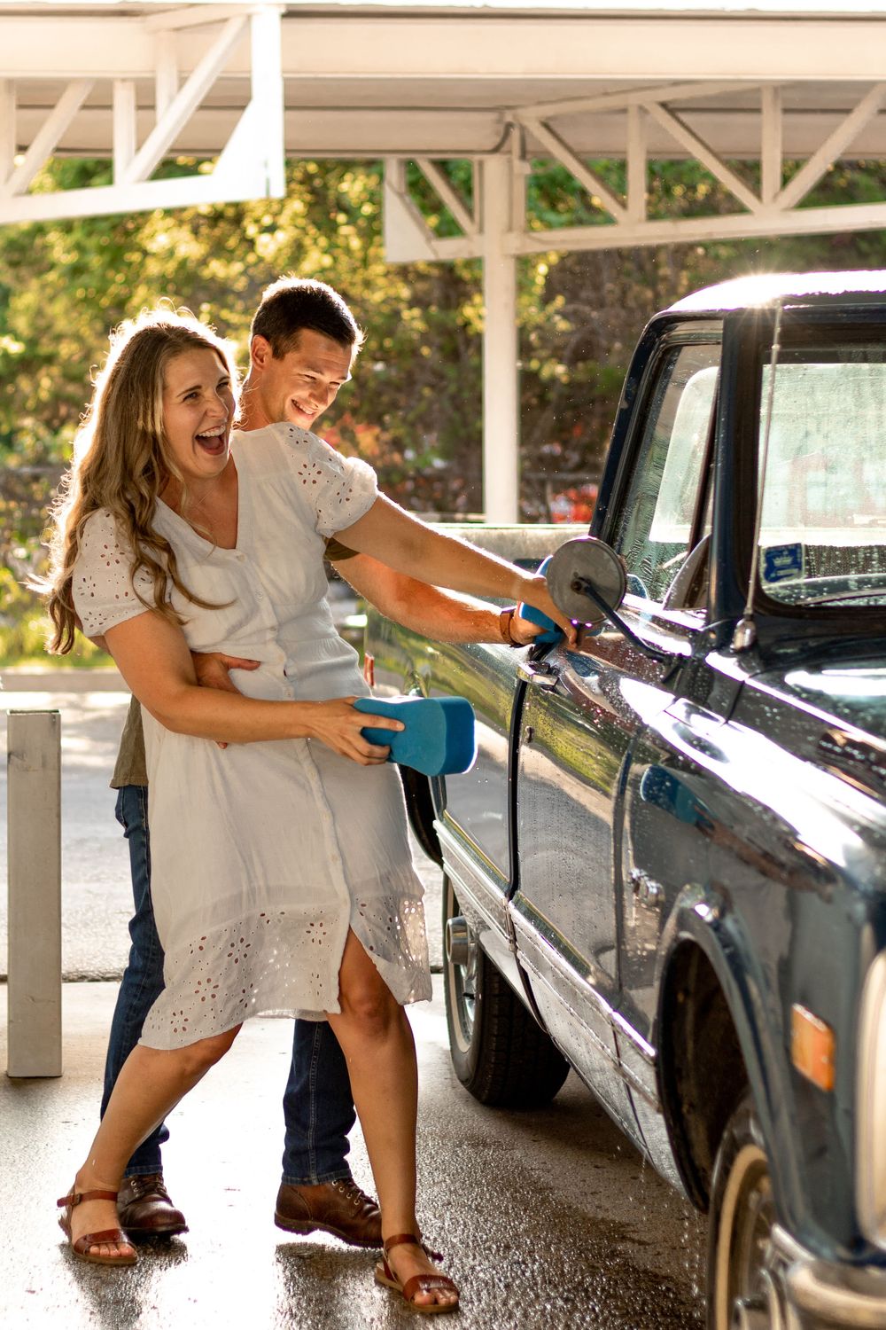 Playful couple washing their vintage truck under a covered parking area.