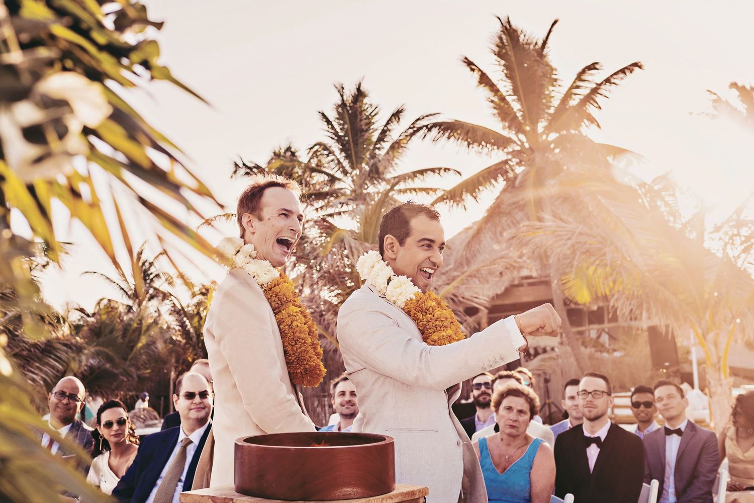 Two people in white outfits perform a traditional ceremony outdoors with palm trees and guests at sunset.
