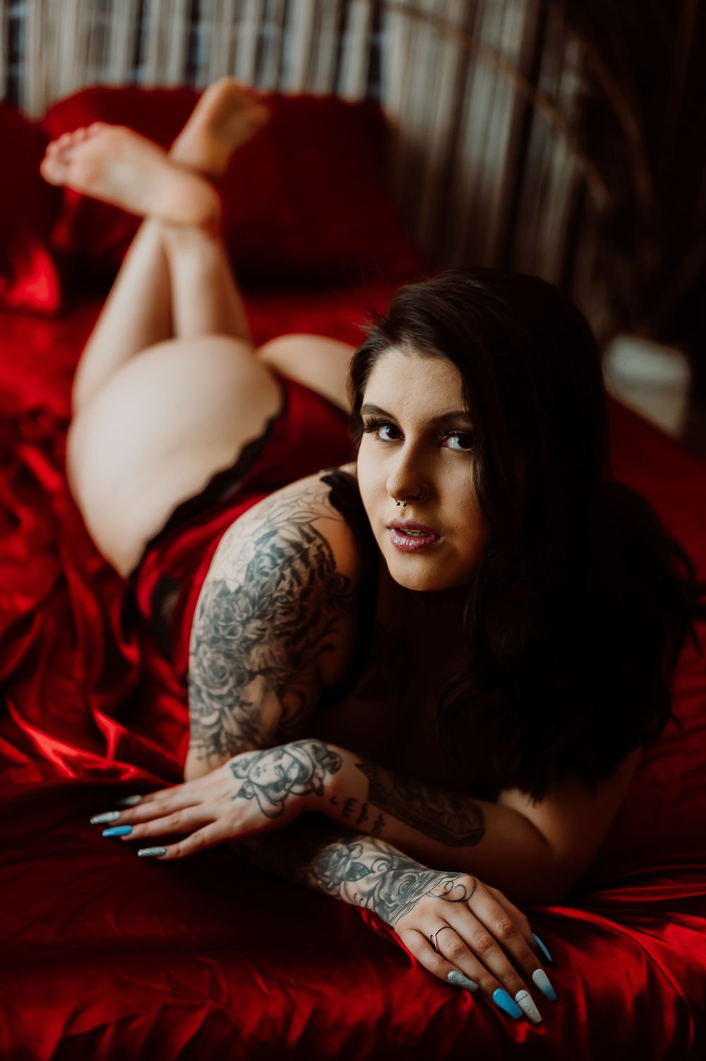 Moody boudoir photography with dramatic lighting showcasing tattoo art against red bedding.