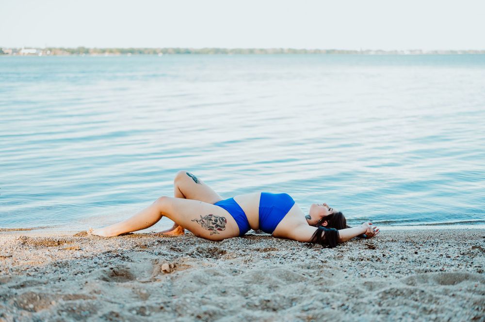 Woman in blue swimsuit lying on sandy beach near calm waters during sunset.