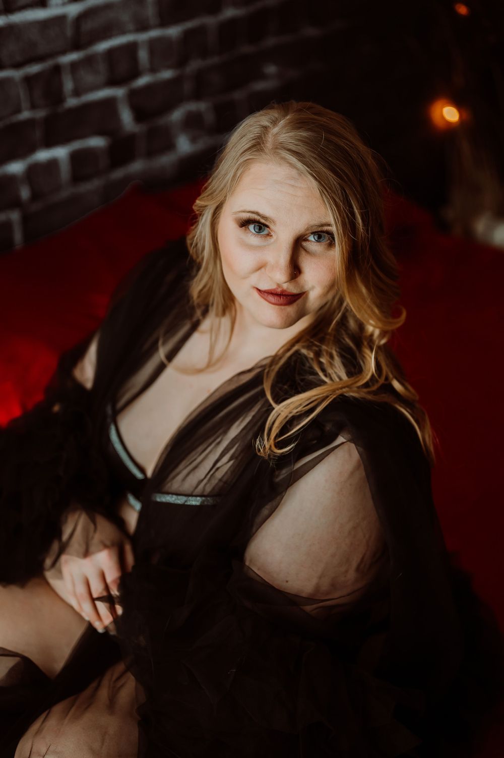 Dramatic boudoir portrait in black lace against brick wall with moody lighting.