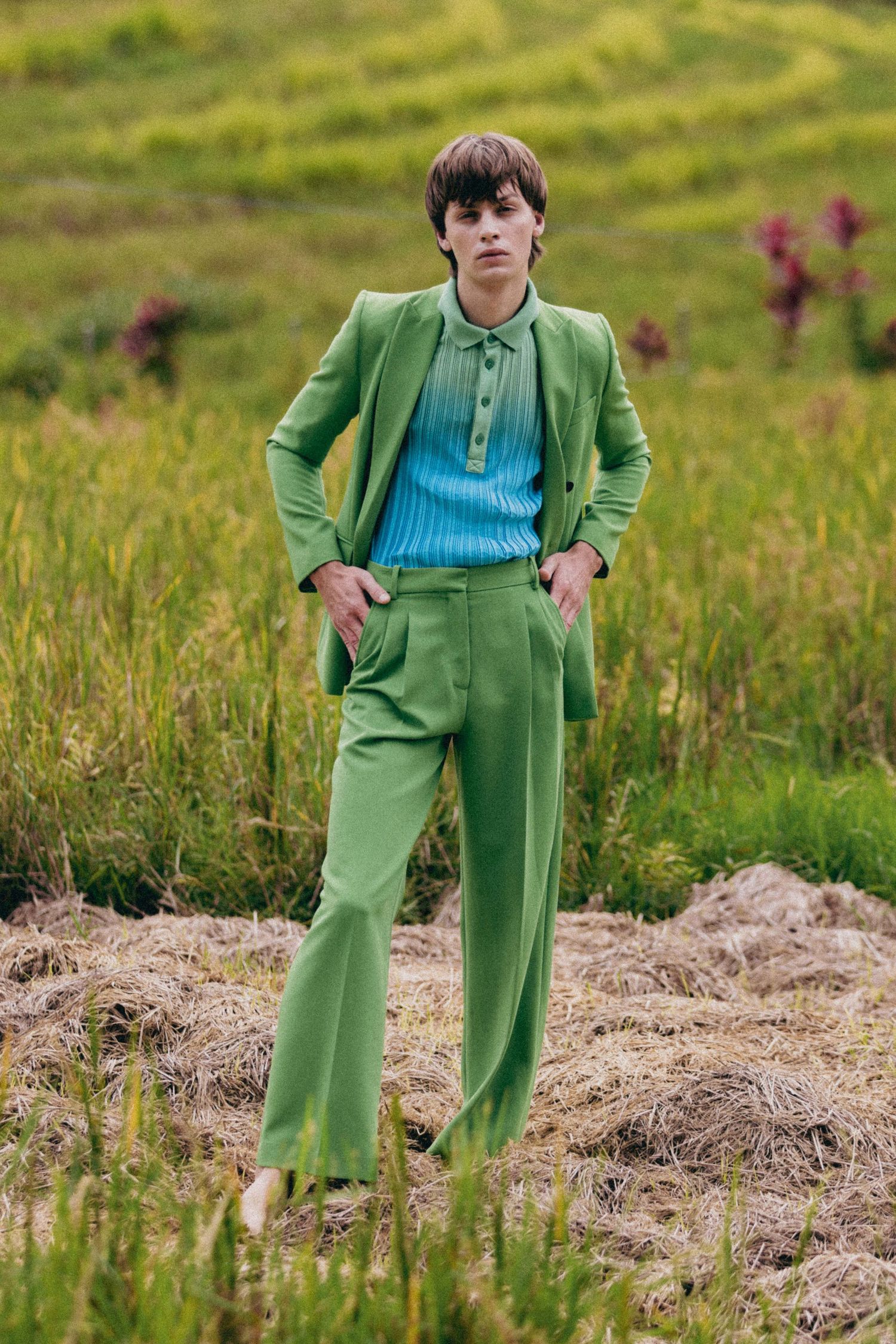 Fashion model wearing bright green suit and blue sweater poses in grassy field.