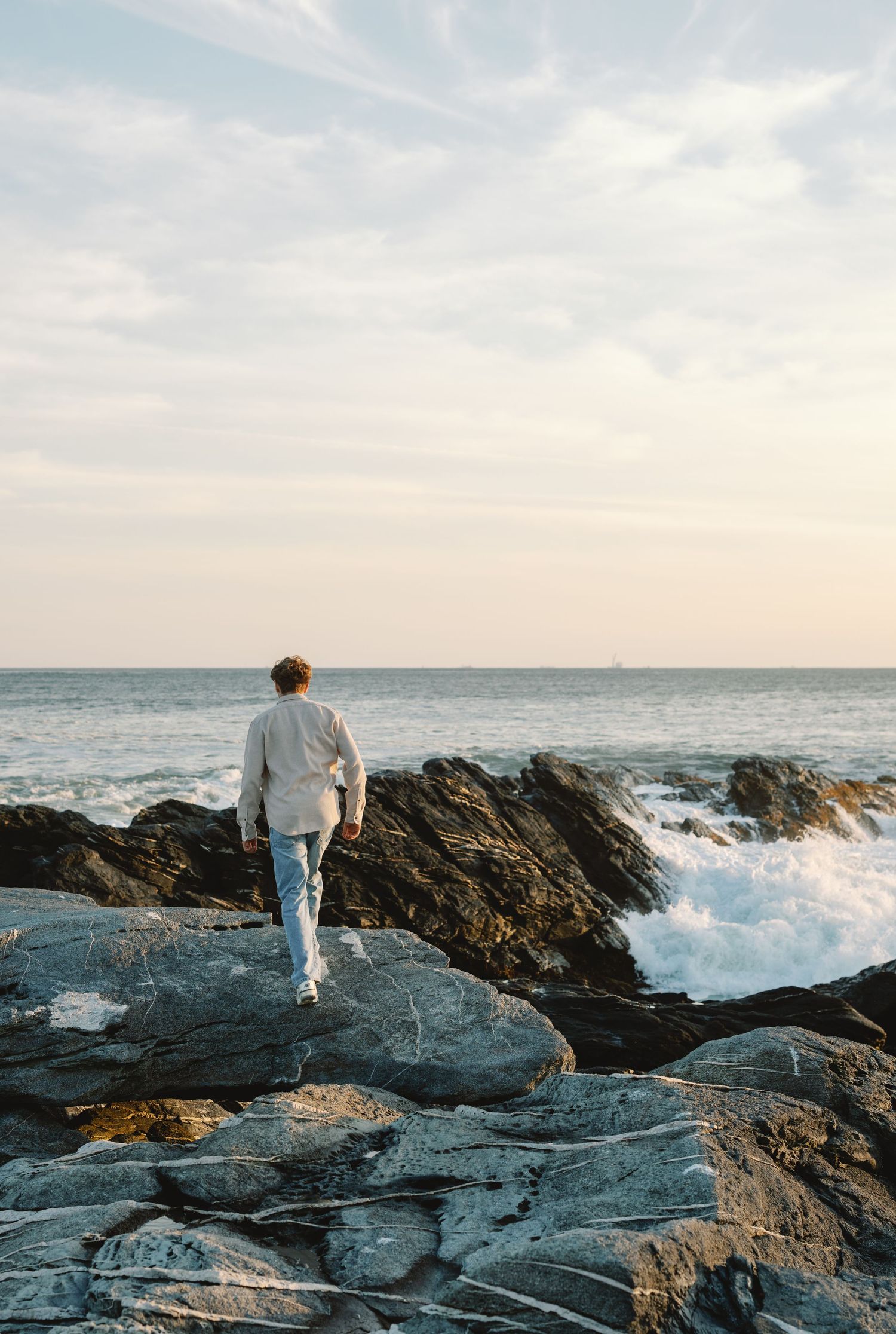 A person in a blue shirt and khaki shorts stands on coastal rocks overlooking the ocean.
