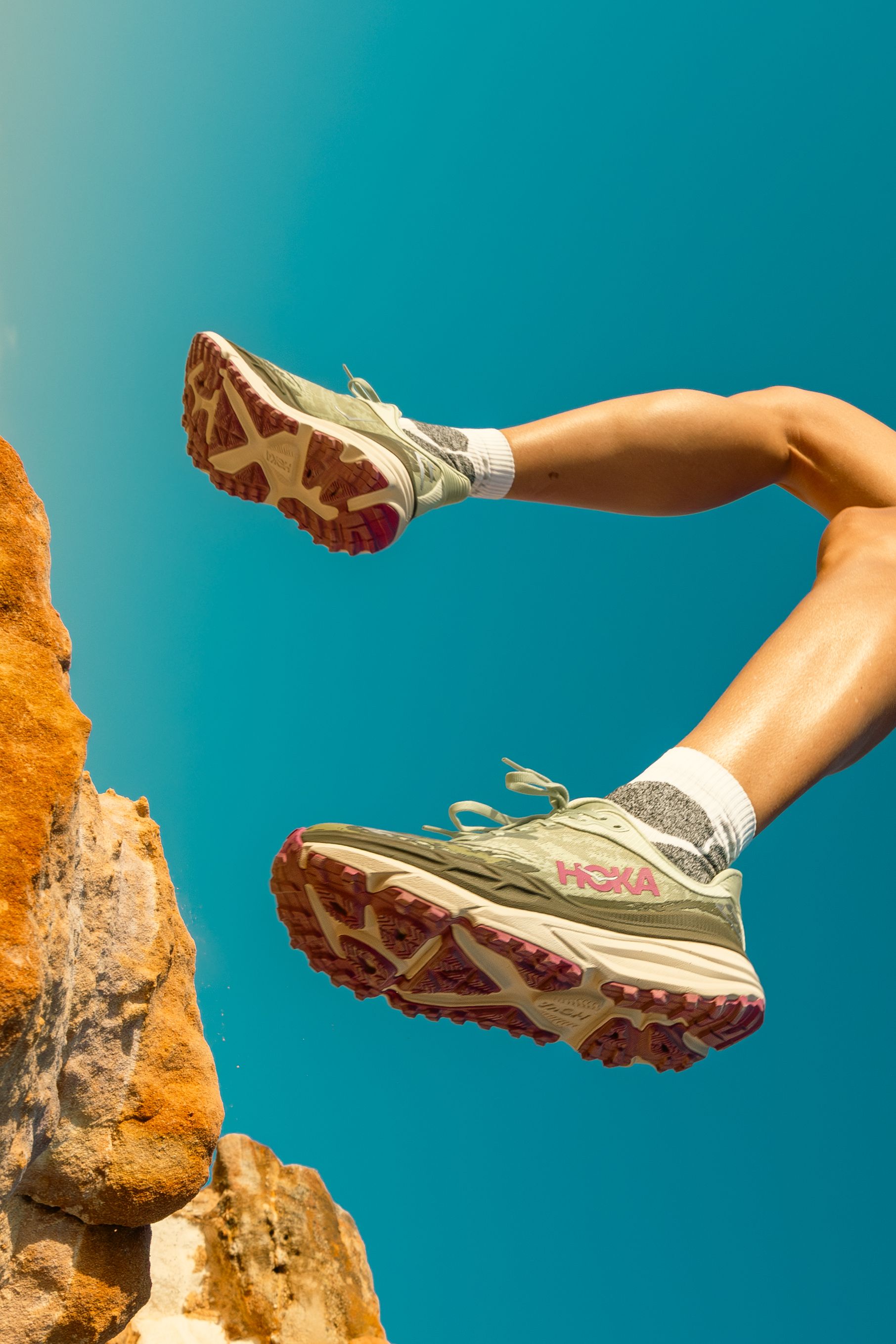 Running shoes with rugged soles dangling over orange cliffs against vivid turquoise sky.