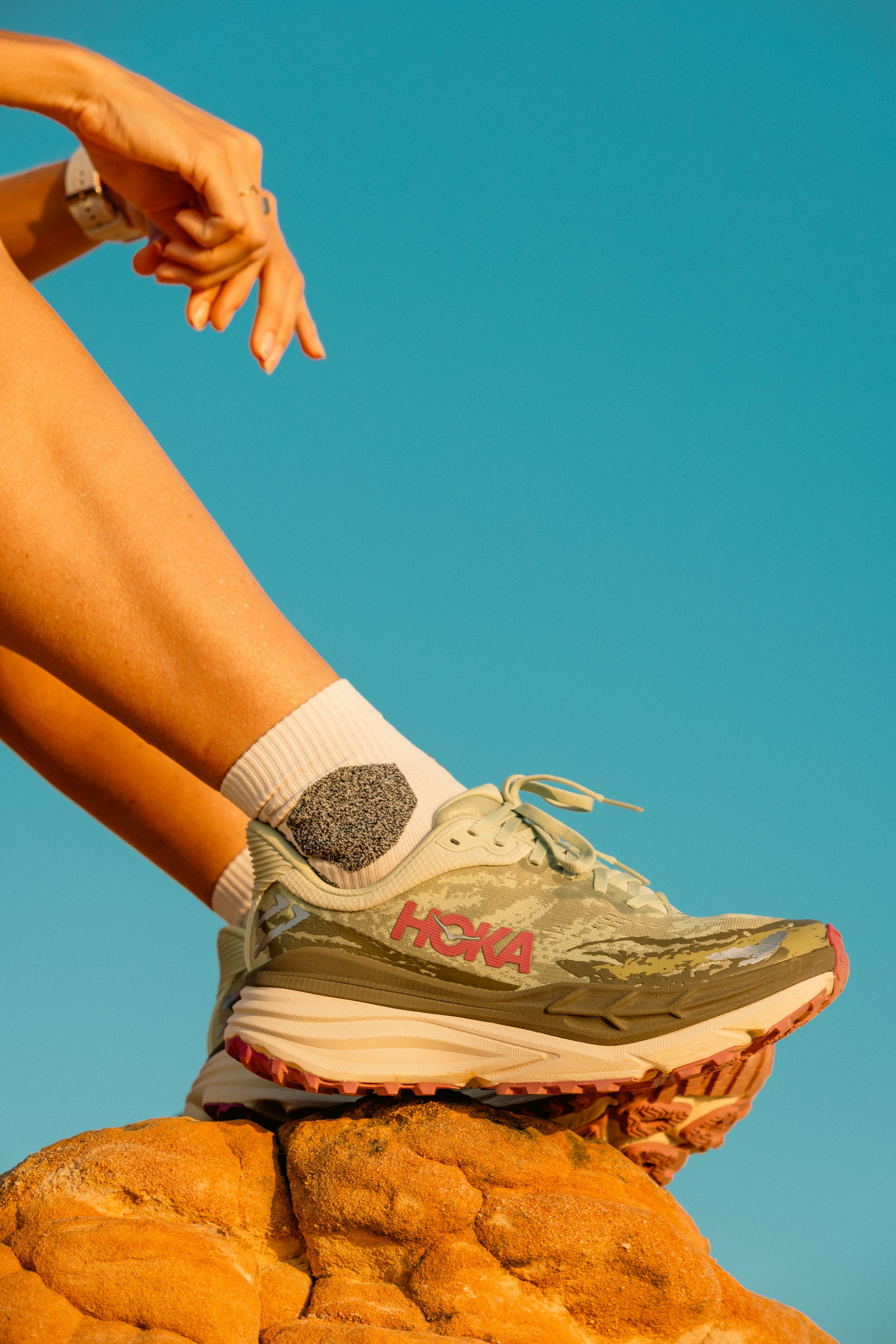 Hoka running shoe perched on orange rock formation against turquoise sky.