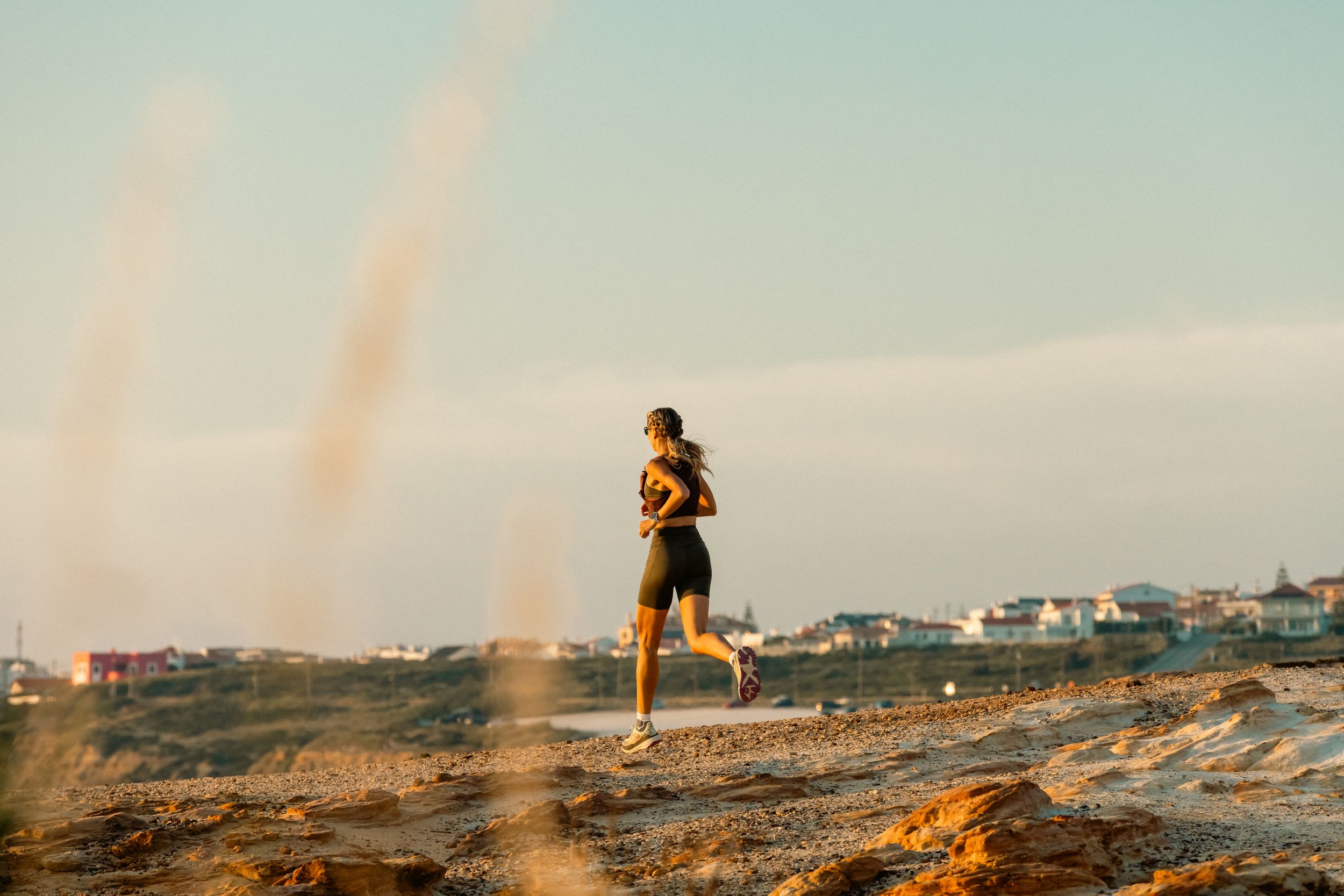 A runner jogs along a scenic coastal path during golden hour with buildings visible in the distance.