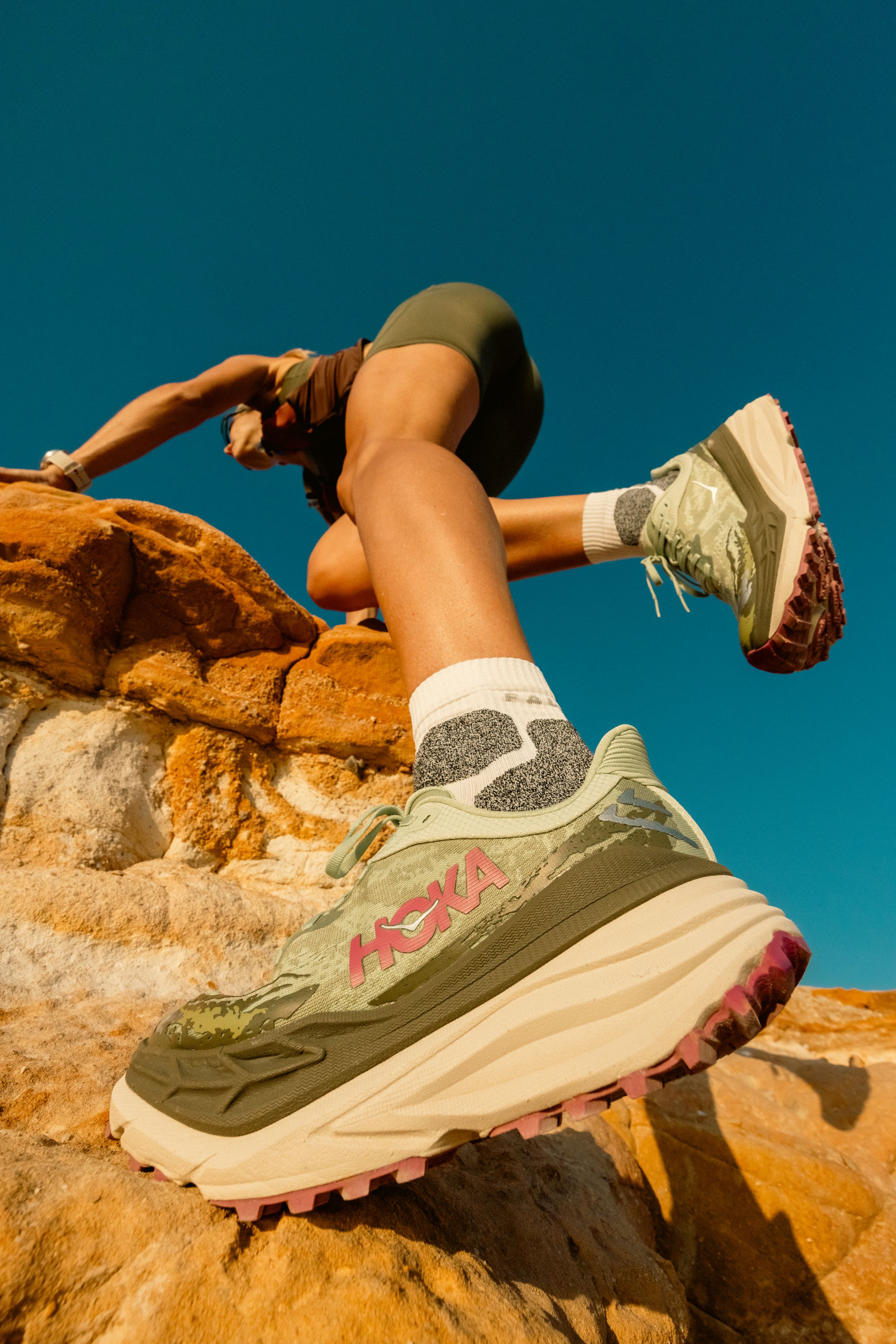 Close-up of Hoka running shoe on rock formation with bright blue sky backdrop.