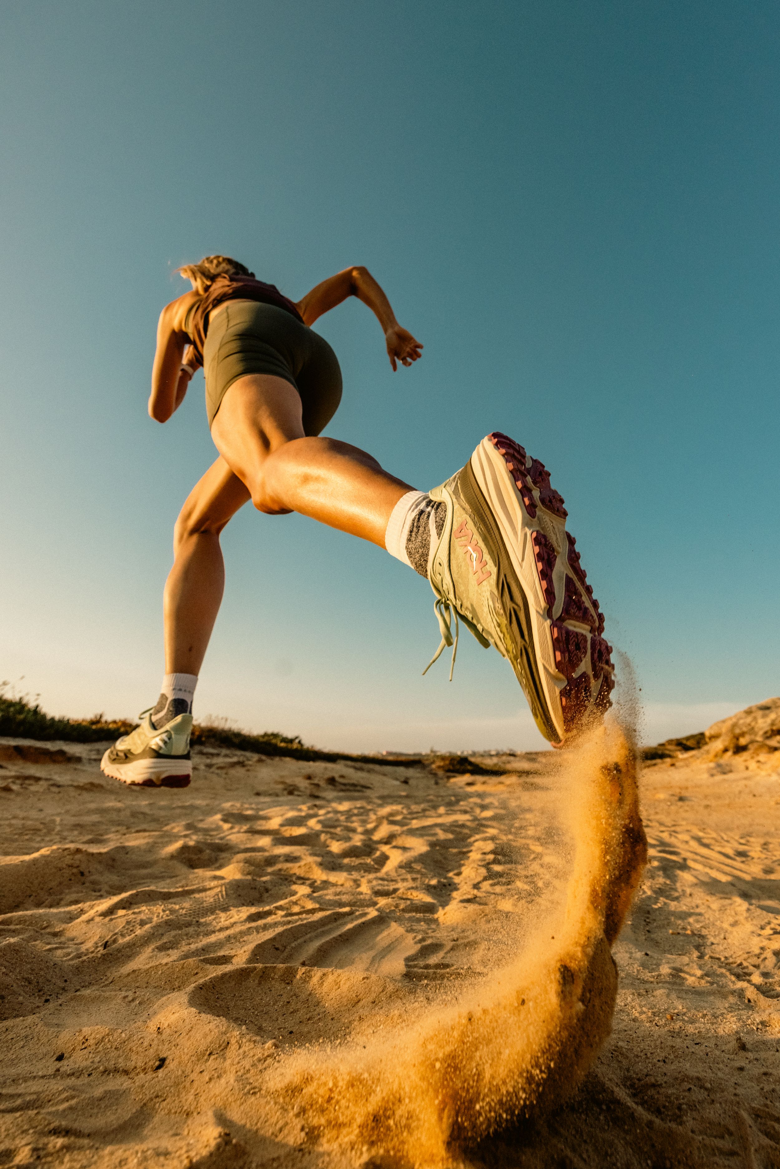 Runner kicking up sand on desert trail wearing Hoka shoes in golden sunset light.