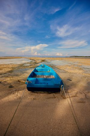 Blue wooden boat stranded on sandy beach during low tide under cloudy sky.
