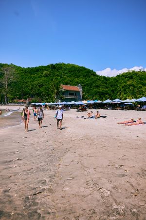 Beachgoers walking along sandy shore with umbrellas and mountains in background.