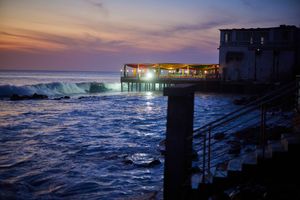 Illuminated oceanfront pier restaurant at dusk with waves crashing below.