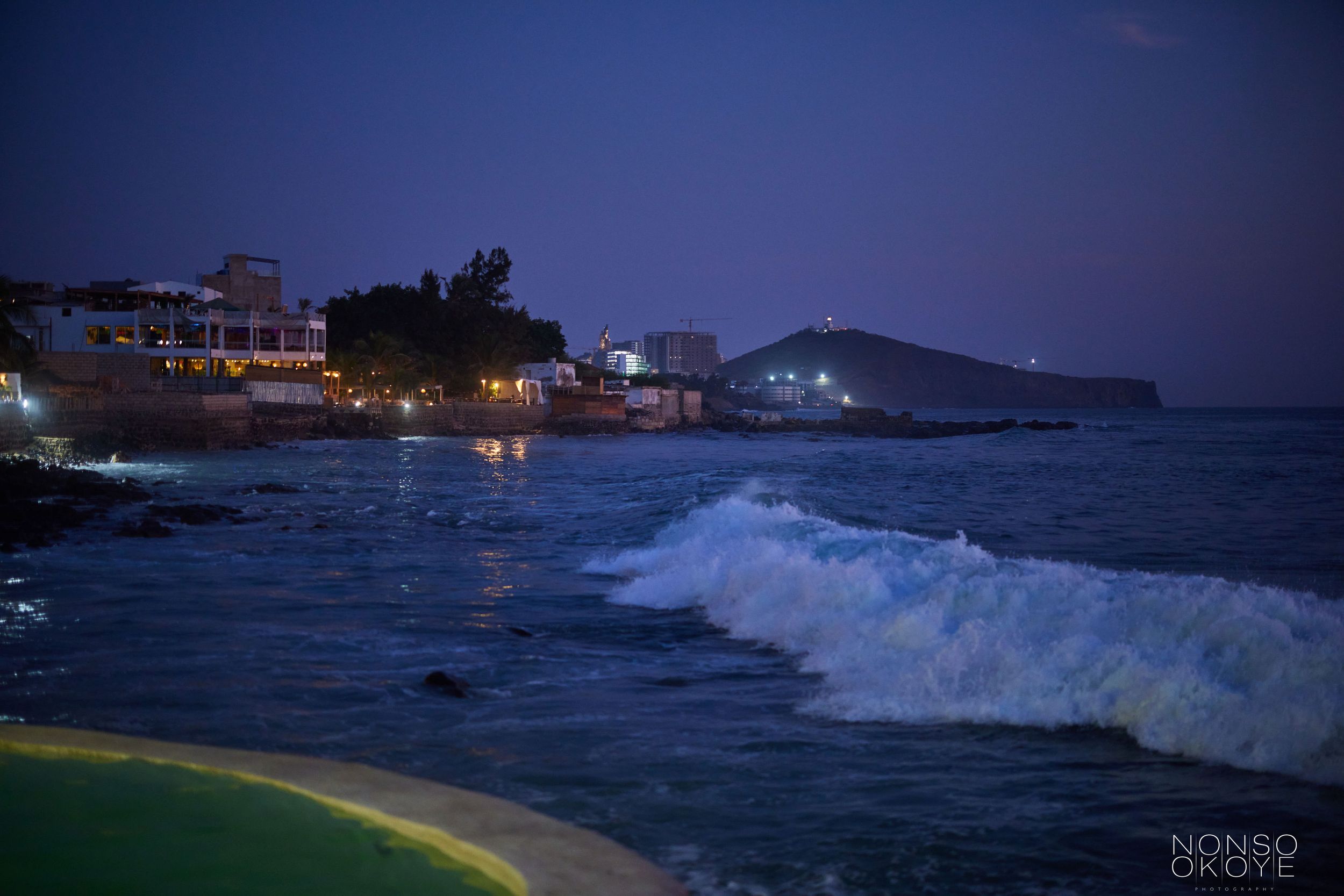 Nighttime coastal scene with crashing waves and illuminated buildings along the shoreline.