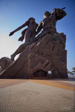 Monumental stone sculpture depicting figures reaching skyward against blue sky.