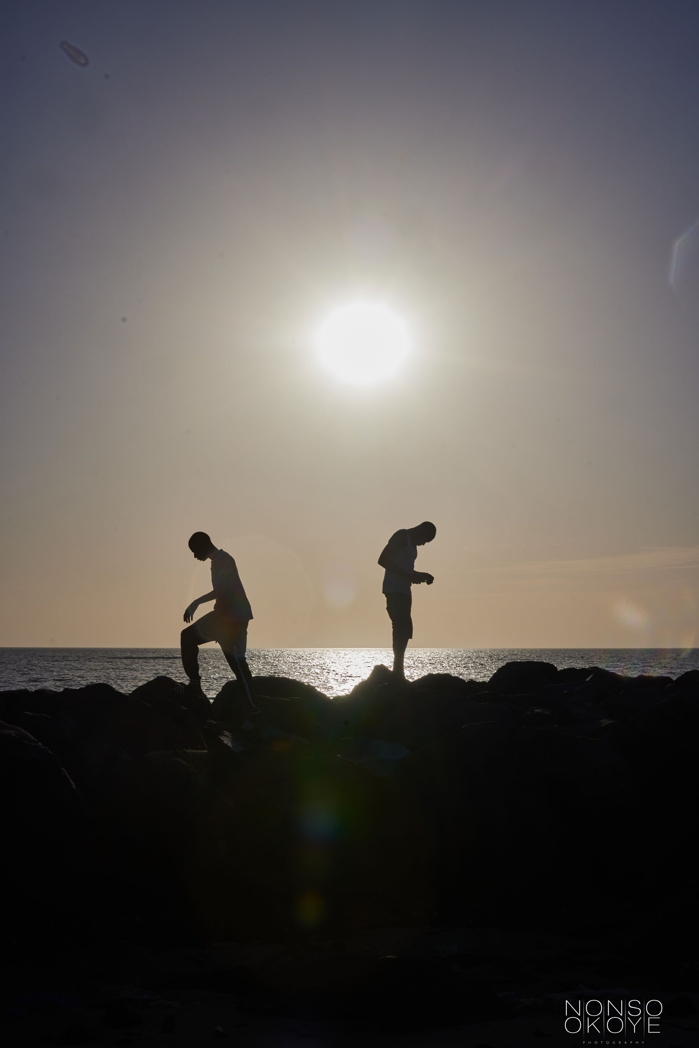 Two silhouettes leaping on coastal rocks against dramatic sunset and ocean backdrop.