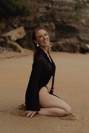Beach photoshoot with person in black outfit posing on golden sand against rocky cliffs.