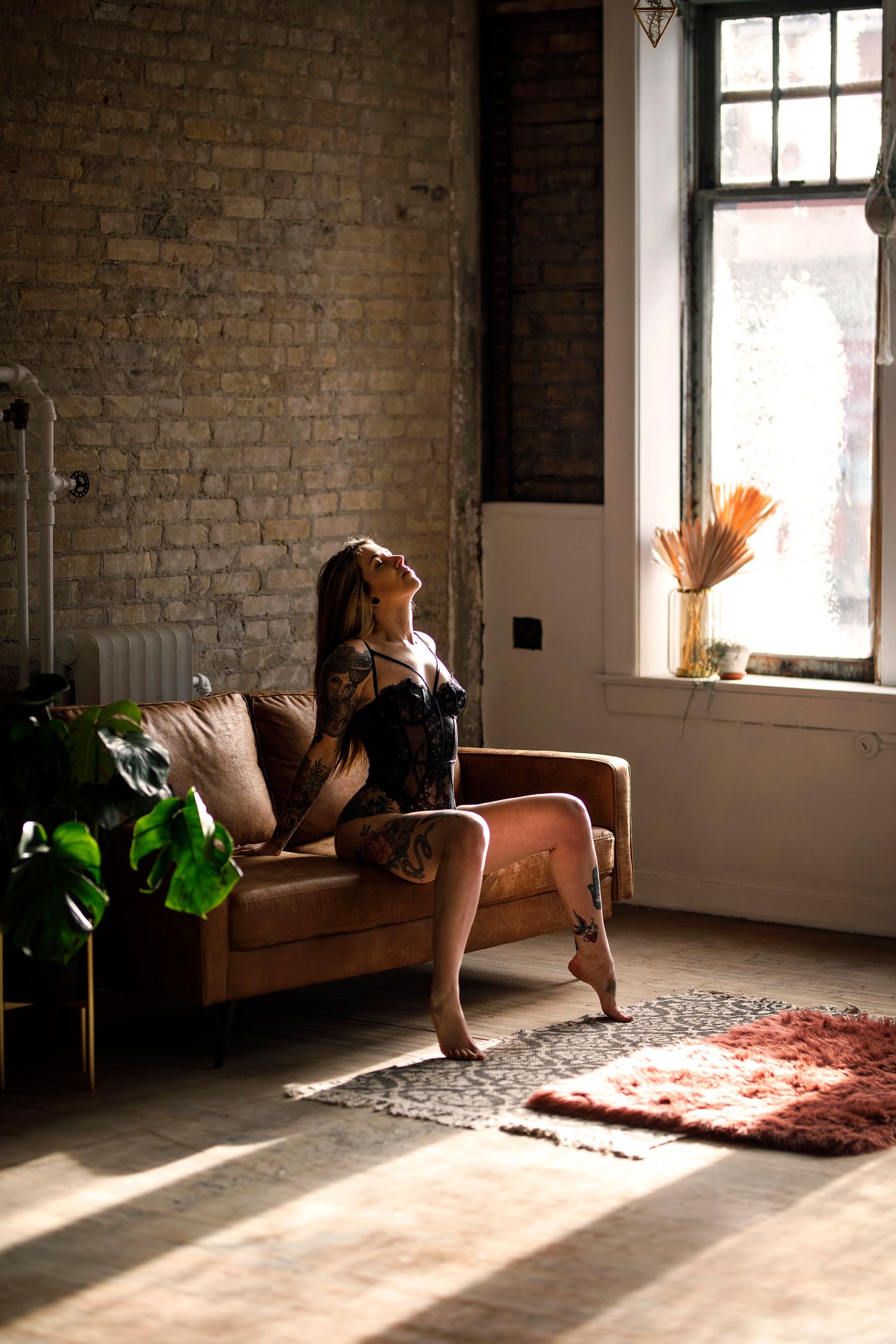 Woman in black lingerie posing on a brown velvet sofa in a sunlit loft with exposed brick walls.