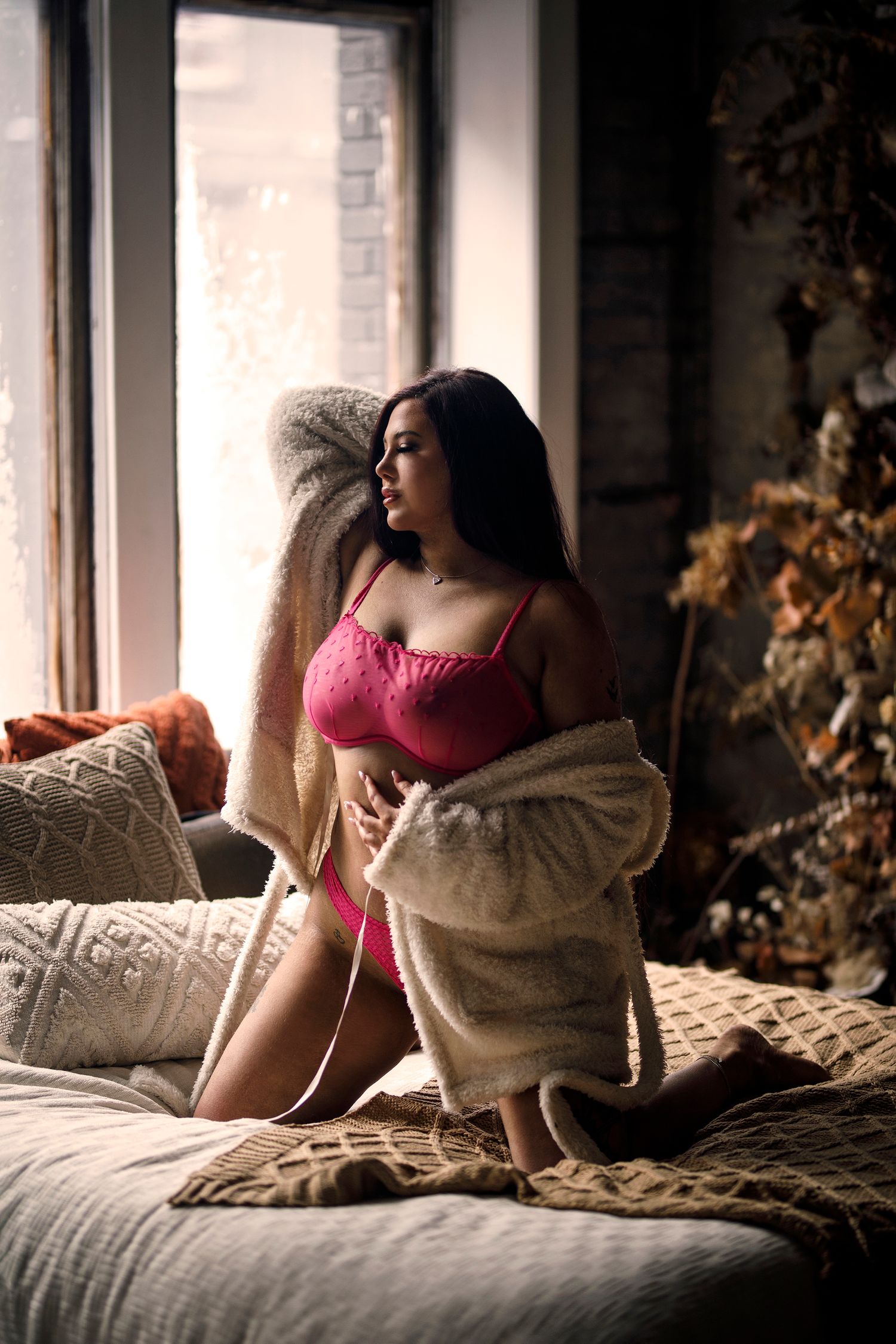 Woman in pink lingerie wrapped in cozy blanket sitting on bed near window in moody interior setting.