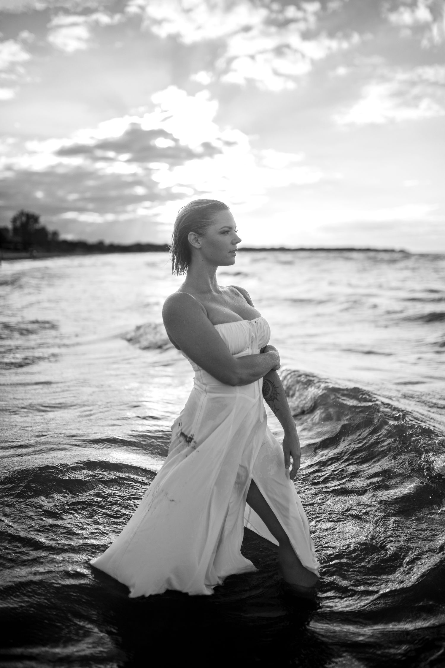 Black and white photo of a woman in a white dress standing in ocean waves at sunset.