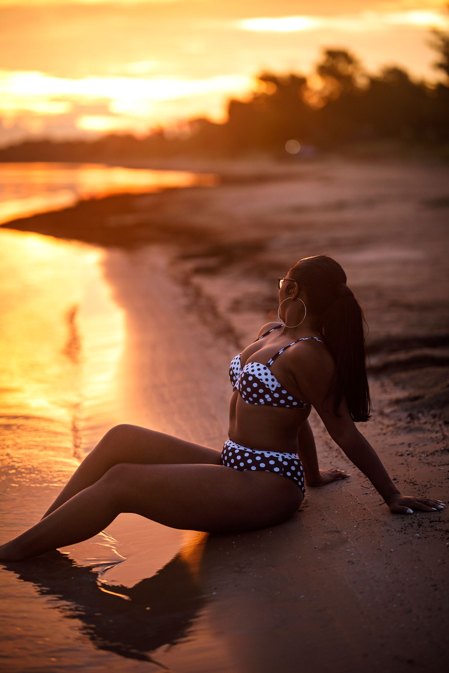 Woman in polka dot bikini sitting on sandy beach at golden sunset, silhouetted against vibrant orange sky.