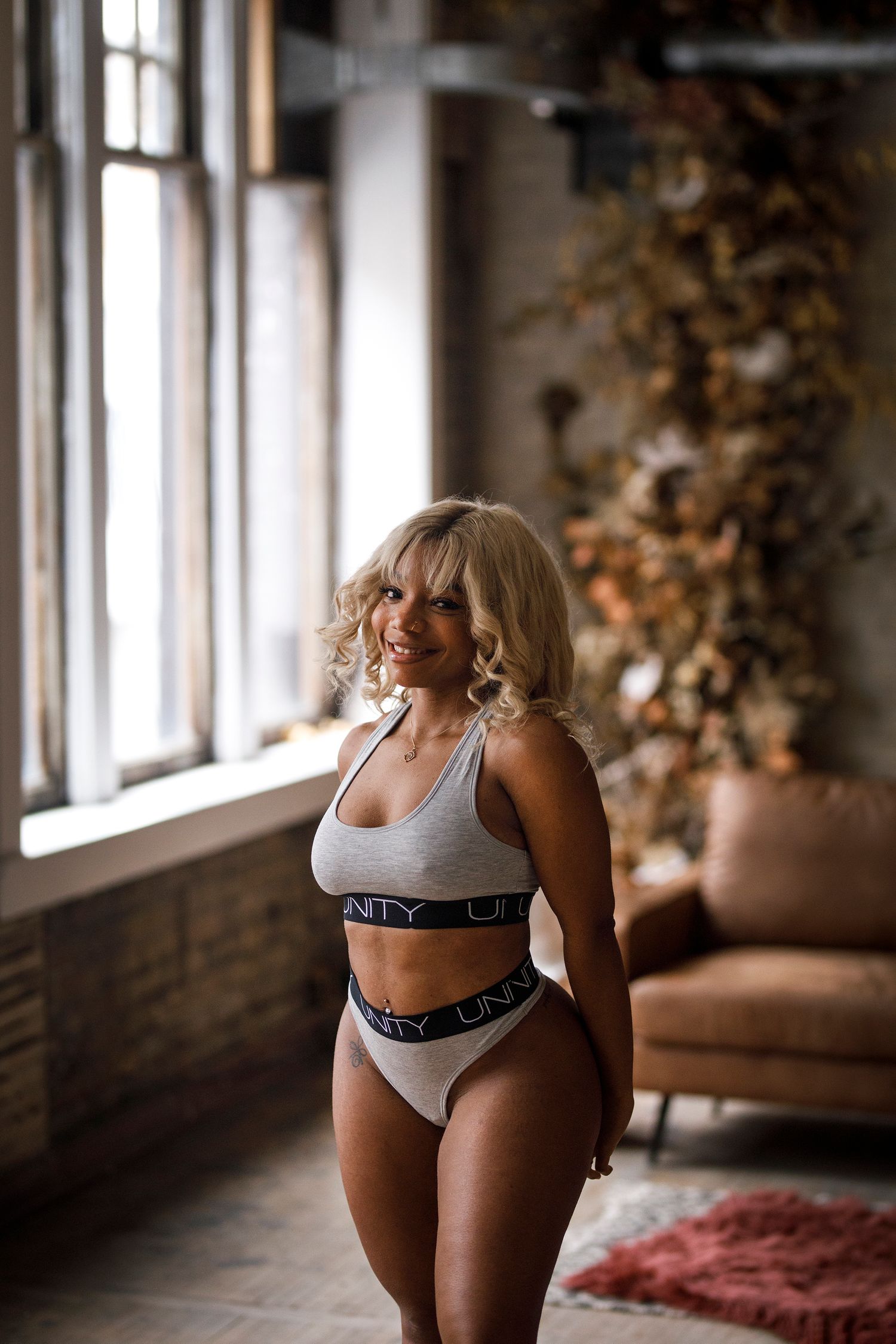 Smiling blonde woman posing in gray Unity branded sports bra and underwear set in a stylish loft interior.